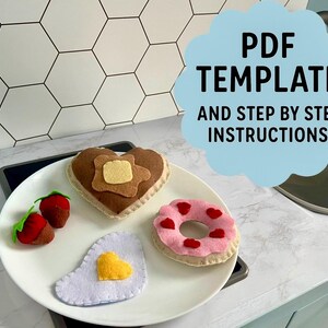 May include: A white plate holds felt food items: a heart-shaped pancake with butter, a pink donut with red hearts, two strawberries, and a fried egg. The image includes text that says "PDF TEMPLATE AND STEP BY STEP INSTRUCTIONS".