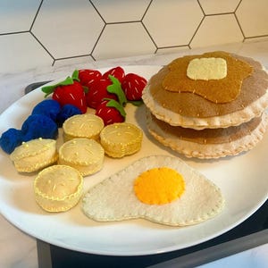 May include: A felt breakfast set on a white plate. The set includes a stack of pancakes with a butter pat, an egg, strawberries, blueberries, and biscuits. The felt food is arranged on a white plate.