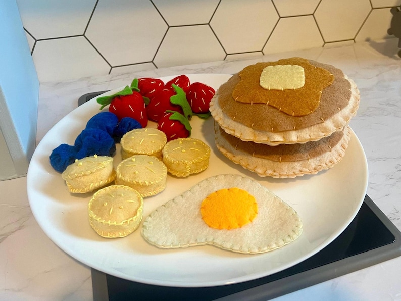 May include: A felt breakfast set on a white plate. The set includes a stack of pancakes with a butter pat, an egg, strawberries, blueberries, and biscuits. The felt food is arranged on a white plate.