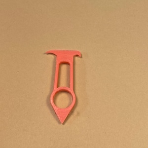 May include: A coral-colored, 3D-printed tool with a unique design. It features a ring at the bottom, a central handle, and a curved top. The tool's pointed end and overall shape suggest a specific function, possibly for crafts or DIY projects.