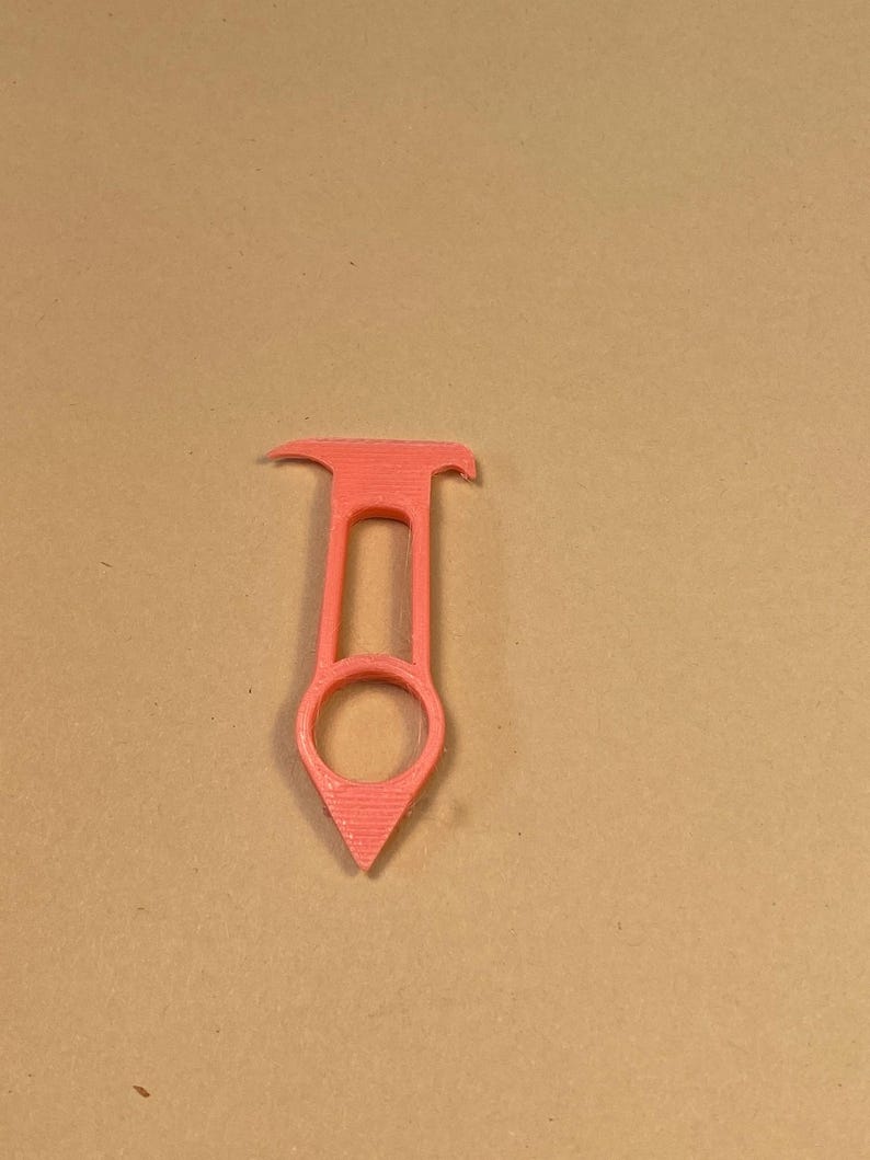 May include: A coral-colored, 3D-printed tool with a unique design. It features a ring at the bottom, a central handle, and a curved top. The tool's pointed end and overall shape suggest a specific function, possibly for crafts or DIY projects.