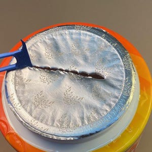 May include: Close-up of a container with a silver foil seal, partially opened with a blue plastic tool. The container has an orange and yellow label. The foil seal has a floral pattern.