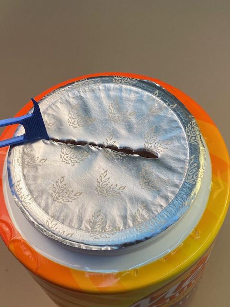 May include: Close-up of a container with a silver foil seal, partially opened with a blue plastic tool. The container has an orange and yellow label. The foil seal has a floral pattern.