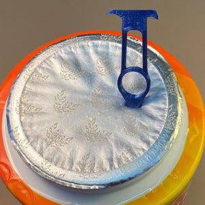 May include: A blue plastic lid lifter is attached to the foil seal of a container. The container has an orange, yellow, and white label. The foil seal has a floral pattern.