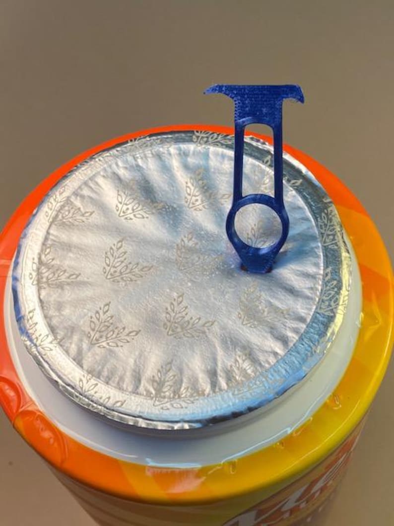 May include: A blue plastic lid lifter is attached to the foil seal of a container. The container has an orange, yellow, and white label. The foil seal has a floral pattern.