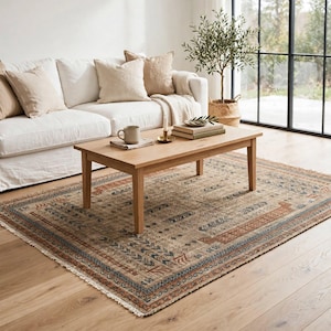 Puede incluir: Una alfombra rectangular con base beige y patrones geom&eacute;tricos intrincados en tonos naranja &oacute;xido, azul pizarra y crema. La alfombra tiene flecos cortos en los bordes y est&aacute; colocada sobre un suelo de madera clara bajo una mesa de centro de madera.