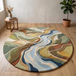 Puede incluir: Una alfombra redonda con un diseño de paisaje texturizado de varias capas con formas orgánicas fluidas en tonos verde salvia, amarillo mostaza, terracota, crema y varios tonos de azul, creando un patrón topográfico similar a un río.