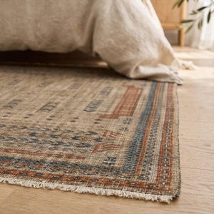 Puede incluir: Una alfombra rectangular con base color canela y patrones geom&eacute;tricos intrincados en tonos naranja &oacute;xido, azul marino y crema. La alfombra presenta un borde decorativo y flecos cortos en el extremo, con una textura tejida en toda la pieza.
