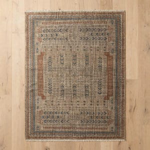 Puede incluir: Una alfombra rectangular con una base de color tostado desgastado y patrones geom&eacute;tricos intrincados en azul apagado y naranja &oacute;xido. El dise&ntilde;o incluye un motivo central, un borde detallado y flecos cortos en los bordes superior e inferior.