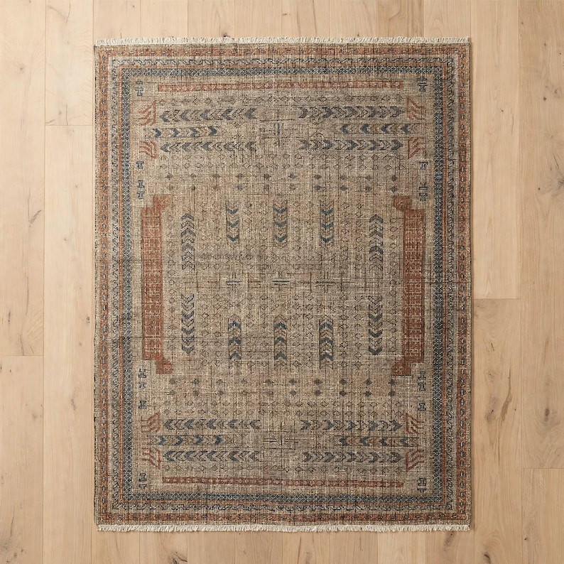 Puede incluir: Una alfombra rectangular con una base de color tostado desgastado y patrones geom&eacute;tricos intrincados en azul apagado y naranja &oacute;xido. El dise&ntilde;o incluye un motivo central, un borde detallado y flecos cortos en los bordes superior e inferior.