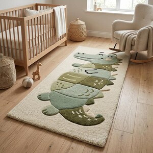 Puede incluir: Una alfombra rectangular que presenta un gran gráfico de cocodrilo estilizado en tonos verde salvia, oliva y bosque. El cocodrilo se muestra con un diseño segmentado y lúdico sobre un fondo color crema con una superficie de pelo suave y texturizado.