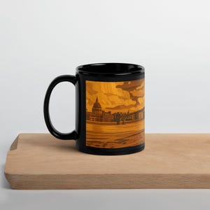 Può includere: Una tazza in ceramica nera con una dettagliata illustrazione di un paesaggio urbano dai toni caldi. L'opera raffigura edifici, una cupola e un fiume sotto un cielo nuvoloso. La tazza è appoggiata su una superficie di legno.