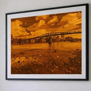 Puede incluir: Obra de arte enmarcada que representa un paisaje urbano con un puente, edificios y un río, renderizado en tonos cálidos y terrosos. La obra de arte está enmarcada en un marco negro y montada en una pared blanca. El estilo parece ser una xilografía o marquetería.