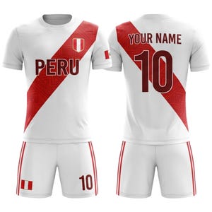 Peut inclure: Uniformes de football blancs avec une bande diagonale et des accents rouges. Le maillot affiche le mot "PERU" et un écusson sur le devant, avec le numéro "10" et "YOUR NAME" au dos. Un short blanc assorti avec des rayures rouges et le numéro "10" complètent l'ensemble.