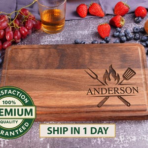 May include: A rectangular wooden cutting board with a carved design featuring a fork, spatula, and flames, with the name "ANDERSON" below. The board is surrounded by grapes, strawberries, blueberries, and kiwi.