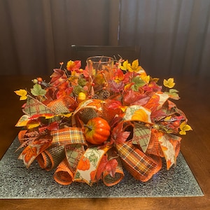May include: A fall-themed centerpiece with orange, green, and brown plaid ribbon, faux leaves, and a small orange pumpkin. The centerpiece is on a glass table.