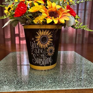 May include: A yellow metal planter with a black chalkboard design featuring sunflowers and the text "You are my sunshine". The planter is filled with a bouquet of yellow sunflowers and red flowers.