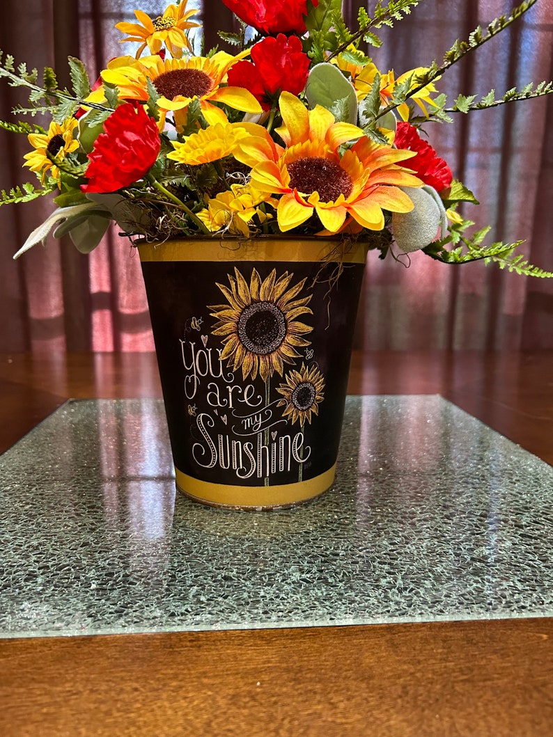 May include: A yellow metal planter with a black chalkboard design featuring sunflowers and the text "You are my sunshine". The planter is filled with a bouquet of yellow sunflowers and red flowers.