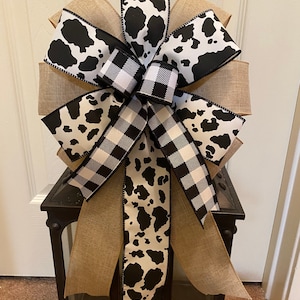 May include: A black and white cow print ribbon bow with a black and white buffalo plaid ribbon bow on top. The bow is tied around a burlap ribbon and is attached to a black lantern.