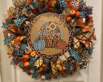 May include: A fall-themed wreath with a brown, leopard print background and the text "Hello Fall". The wreath is decorated with orange, blue, and brown ribbons, as well as small pumpkins and sunflowers.