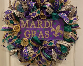 Puede incluir: Una corona de Mardi Gras con cintas y malla de color morado, dorado y verde. La corona tiene un letrero morado con letras doradas brillantes que dicen "Mardi Gras".