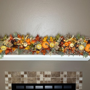 May include: A long, horizontal autumn-themed garland featuring artificial orange pumpkins, yellow and red maple leaves, green foliage, and small berry clusters. The decorative arrangement is displayed across a white mantelpiece shelf.