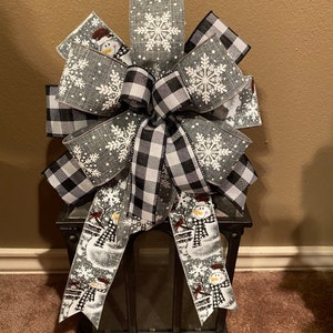 May include: A black and white buffalo plaid ribbon bow with a gray ribbon with a snowflake pattern and snowman design. The bow is tied around a black lantern.