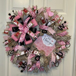 May include: A pink and brown Valentine's Day wreath with a burlap base, pink and white ribbon, and a pink envelope with the text "P.S. I love you".