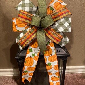 May include: A green, white, and orange plaid bow with a pumpkin print ribbon tied around a black metal lantern.