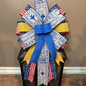 May include: A decorative bow in patriotic colors, featuring blue, red, and yellow ribbons. The ribbons are printed with text such as "Air Force", "Freedom", and "Hero". The bow is placed on top of a black lantern.