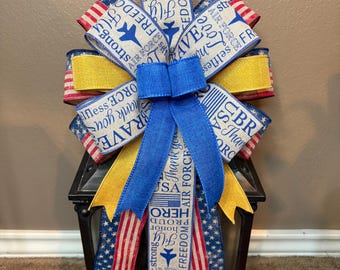 May include: A decorative bow in patriotic colors, featuring blue, red, and yellow ribbons. The ribbons are printed with text such as "Air Force", "Freedom", and "Hero". The bow is placed on top of a black lantern.