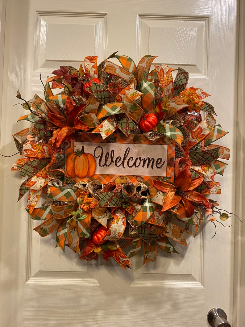 K&ouml;nnte beinhalten: Ein Herbstkranz mit orangefarbenen, gr&uuml;nen und braunen B&auml;ndern, Bl&auml;ttern und K&uuml;rbissen. Der Kranz hat ein Schild mit der Aufschrift "Willkommen".
