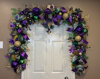 May include: A Mardi Gras door swag with purple, green, and gold ribbon, beads, and ornaments. The swag is decorated with a fleur-de-lis.