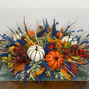 May include: A fall-themed centerpiece featuring white and orange pumpkins, berries, and foliage. The arrangement includes blue accents and is set on a rectangular glass base. The pumpkins have black detailing.