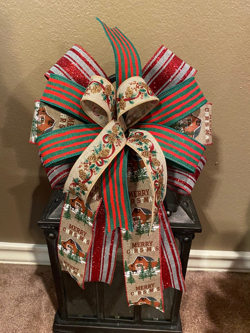 May include: A red, green, and white striped Christmas bow with a burlap ribbon featuring the text "Merry Christmas" and a farmhouse design. The bow is tied around a black lantern.