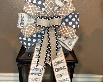 May include: A decorative Easter bow featuring various patterned ribbons. The bow includes ribbons with white polka dots on a gray background, diamond patterns, and bunny silhouettes. The bow is accented with a burlap-style ribbon and long, patterned tails.