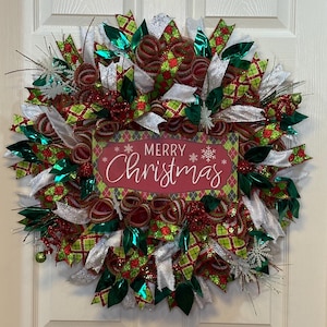 Puede incluir: Una corona navideña festiva con cintas rojas, verdes y blancas, acentuada con purpurina y elementos decorativos. La corona presenta un letrero central que dice "MERRY Christmas" en una fuente cursiva.