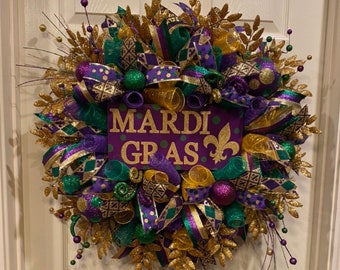 May include: A festive Mardi Gras wreath with purple, green, and gold ribbons and accents. The wreath features a central sign with the words "MARDI GRAS" and a fleur-de-lis symbol. Decorative elements include glittery leaves and beaded sprigs.
