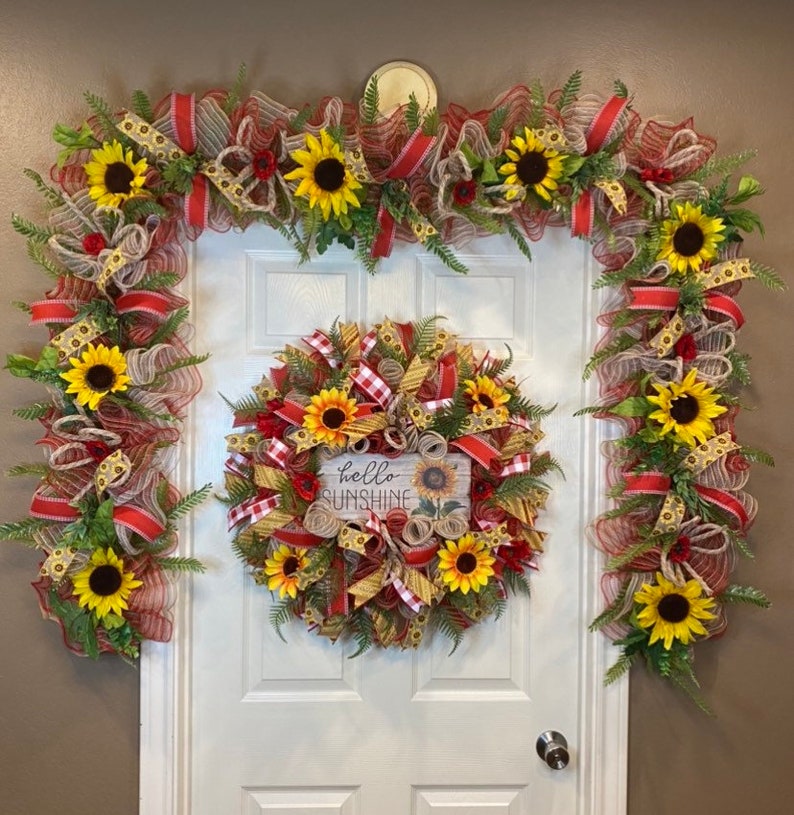 K&ouml;nnte beinhalten: Ein Sonnenblumenkranz mit einem rot-wei&szlig; karierten Band und einem Schild mit der Aufschrift "hello sunshine". Zwei passende Sonnenblumen-Girlanden mit rot-wei&szlig; kariertem Band befinden sich auf beiden Seiten des Kranzes.