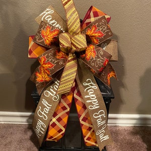 May include: A black lantern with a brown, orange, and red plaid ribbon bow. The bow has a burlap ribbon with the text "Happy Fall Y'all" written on it. The bow is decorated with fall leaves.