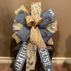 May include: A farmhouse-themed bow with a beige and brown checkered ribbon, a blue ribbon with the text "FARMHOUSE" and a beige ribbon with a chicken print. The bow is tied around a black lantern.
