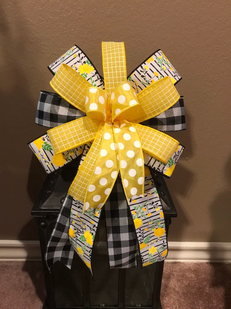 May include: A decorative bow featuring a mix of patterned ribbons. The bow includes yellow polka dots, yellow gingham, black and white gingham, and ribbons with yellow floral and striped designs. The bow is likely for decorating.
