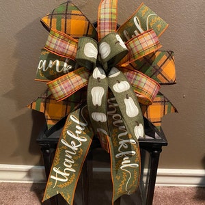 May include: A green and orange plaid bow with a white pumpkin ribbon and the word "thankful" written on it. The bow is tied around a black lantern.