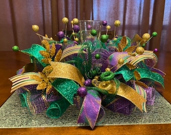 May include: A Mardi Gras themed centerpiece with green, gold, and purple mesh, ribbons, and decorative balls. The centerpiece is on a clear glass base.