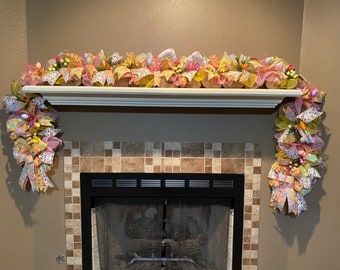 May include: A colorful Easter garland with pink, yellow, and green bows and ribbons, decorated with Easter eggs, bunnies, and carrots. The garland is draped over a white mantel above a fireplace.