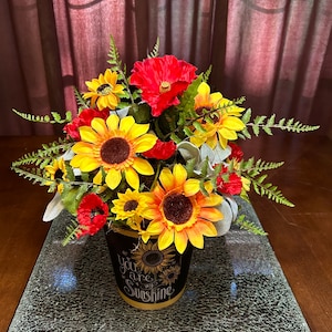May include: A black ceramic flower pot with the text "You are my sunshine" in white lettering. The pot is filled with a floral arrangement of yellow sunflowers, red poppies, and green ferns.