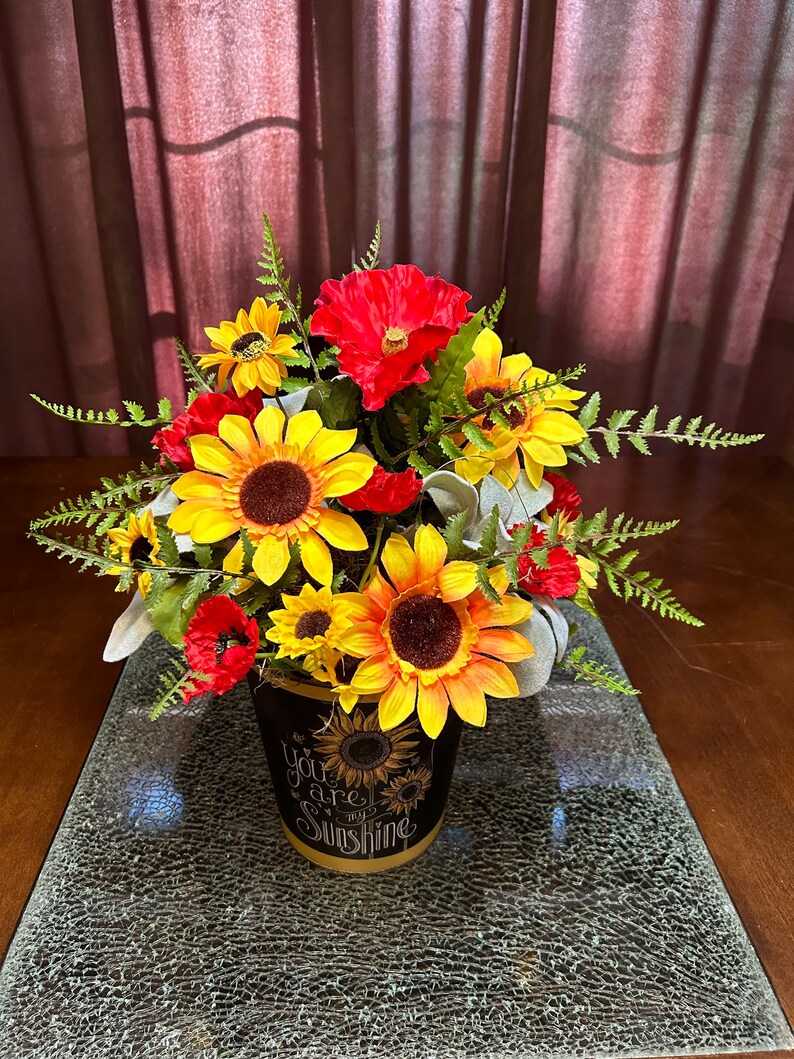 May include: A black ceramic flower pot with the text "You are my sunshine" in white lettering. The pot is filled with a floral arrangement of yellow sunflowers, red poppies, and green ferns.