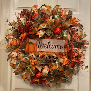 K&ouml;nnte beinhalten: Ein Herbstkranz mit orangefarbenen, gr&uuml;nen und braunen B&auml;ndern, k&uuml;nstlichen K&uuml;rbissen und Bl&auml;ttern. Der Kranz hat ein Schild mit der Aufschrift "Willkommen".