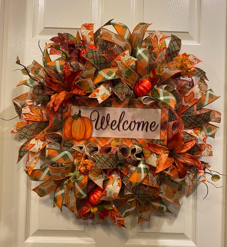 K&ouml;nnte beinhalten: Ein Herbstkranz mit orangefarbenen, gr&uuml;nen und braunen B&auml;ndern, k&uuml;nstlichen K&uuml;rbissen und Bl&auml;ttern. Der Kranz hat ein Schild mit der Aufschrift "Willkommen".