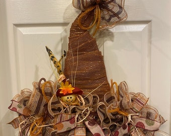 May include: A brown and gold witch hat wreath with a burlap bow and a scarecrow face in the center. The wreath is decorated with brown, gold, and red ribbon.